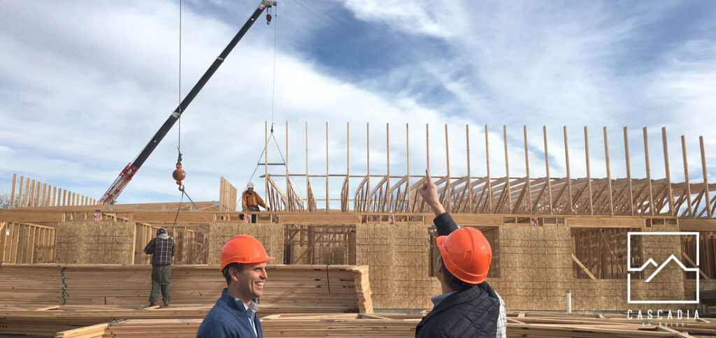 Two men wearing construction hats stand in front of the construction site of a new building. In the background a crane is moving trusses onto the framed walls as two construction workers assist.