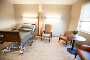 A resident room with a bed, tray, three soft chairs and a small round table sitting in front of a large window.