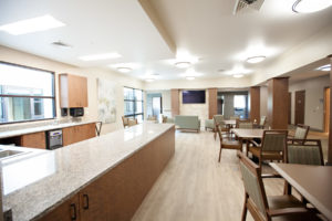 Cafeteria area with a row of square tables with four padded chairs at each table. Across from the tables is a long u-shaped flat counter. At the far end of the room is a love seat and two chairs sitting in front of a well-mounted television.