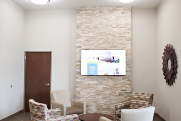 A small sitting area with four soft chairs in a circle and a table in the middle sitting beneath a television mounted to a brick wall.