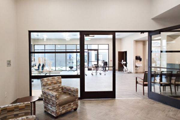 A glass room with two soft chairs and a table placed between them. As you leave the room you enter a room with many types of physical therapy equipment.