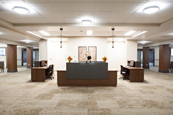 A large work station with a reception desk and two smaller desks on each side. Computers and office chairs are at each desk. Wide hallways can be seen on either side of the work station.