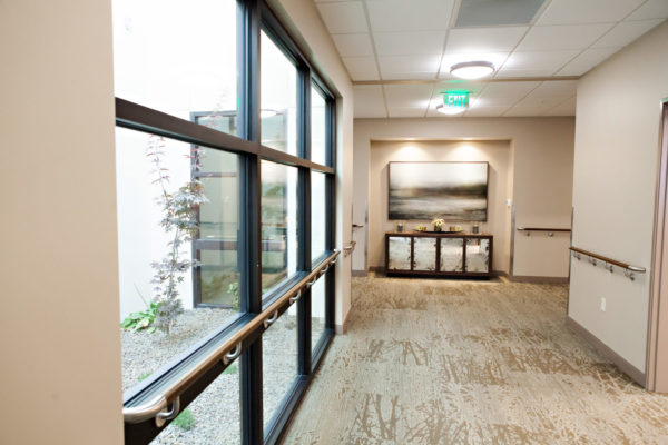 A wide hallway with handrails on either side with large windows on the left. A cabinet and painting sit at the hallway intersection.