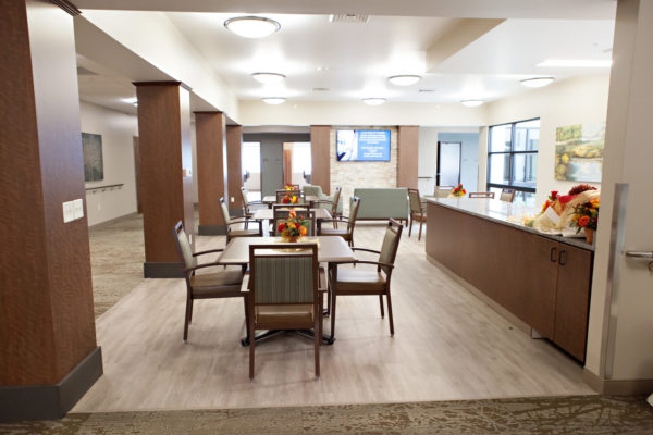 A dining area with four square tables and four chairs at each table. A large counter runs along one side of the room, a love seat and soft chairs are at the far end of the room below a large wall-mounted television.