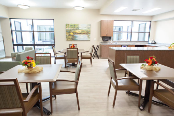 A dining area with square tables and four chairs at each table. A large u-shaped counter is in the corner and a love seat and soft chair are placed in the opposite corner.