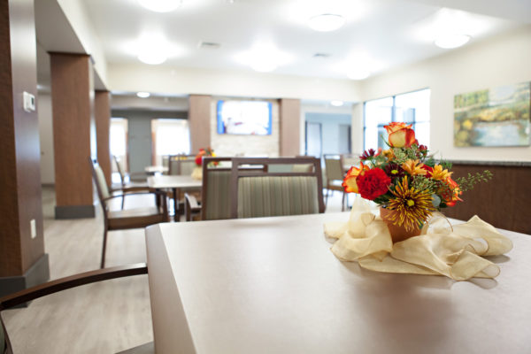 A small clay pot filled with carnations, roses and dahlias sits on a decorative scarf that is placed on a square table with striped padded chairs.