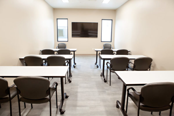 A narrow room with six small rectangular tables and two padded chairs at each table. On the far wall is a large mounted television and two smaller rectangular windows.