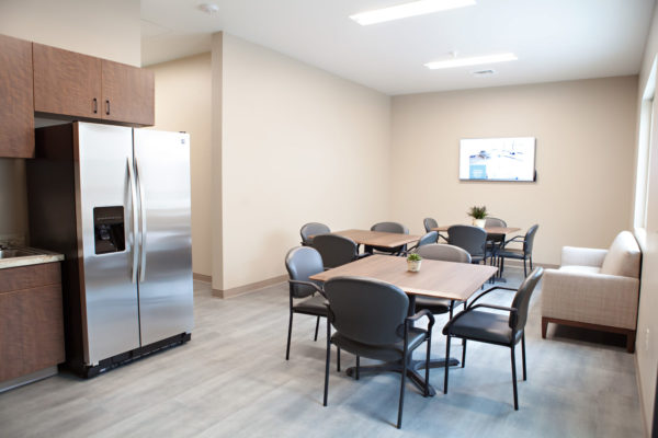 A small room with three square tables and four chairs at each table, and a large side-by side refrigerator and sink. A sofa sits underneath a large window and a television is mounted to the far wall.