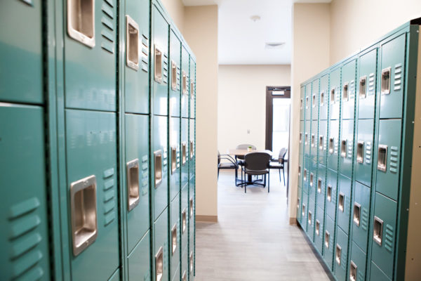 A narrow walkway with four rows of gym-style lockers on each side. The hallway opens into a room with a square table and four chairs.