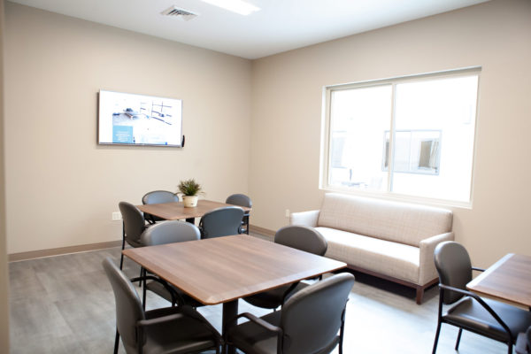 A small room with three square tables and four chairs at each table. A sofa sits underneath a large window and a television is mounted to the far wall.