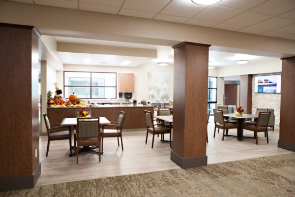 A dining area with square tables and four chairs at each table. In the background is the kitchen area with two large counters, and several large windows.