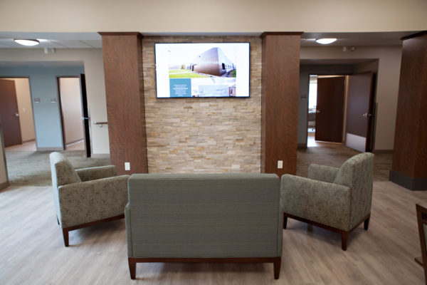 Cascadia of Boise, Idaho lobby. A love seat and two chairs sit in a semi-circle in front of a brick pillared wall with a wall-mounted television. A large hallway leading to resident rooms is on either side of the wall.