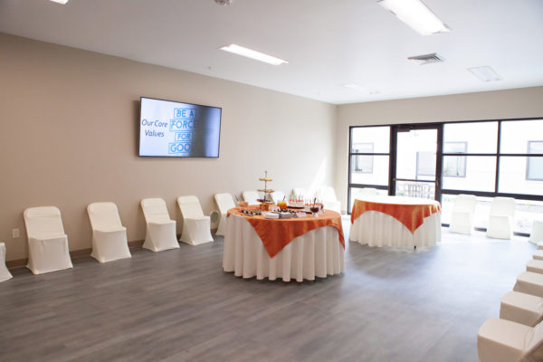 A long room with two large, round tables with tablecloths placed in the center of the room. One table is covered with trays and bowls of desserts. Chairs are evenly spaced around the perimeter of the room including in front of tall window that cover the back wall of the room.