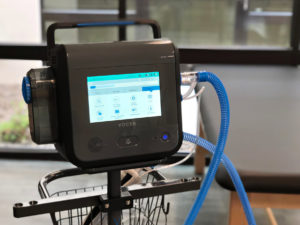 A respiratory ventilator unit on a rolling stand with a digital screen and four tubes connected, a large basket is on the backside of the stand.
