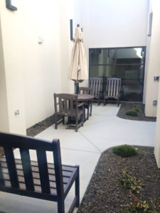 A concrete sidewalk with rocking chairs, a table with an umbrella and chairs, and a bench are placed along the sidewalk. Flower beds are on either side of the sidewalk.