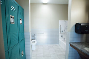 A bathroom used for Occupational Therapy. A tile floored bathroom with two rows of lockers on the left and a sink with a counter across from them. A toilet and tub with grab bars are at the far end of the room.