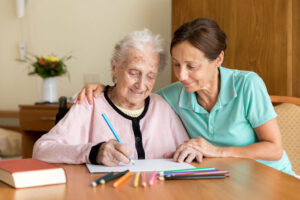 A smiling senior woman is sitting at a desk with a colored pencil in her right hand and a piece of paper in front of her. To her left is a smiling young woman with her right arm around the woman's shoulder and her left hand placed gently on the woman's left hand.