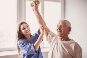 A smiling senior man holding a dumbbell in his right hand with his arm raised overhead is assisted by a smiling young therapist who has her right hand on the man's elbow and her left hand on his wrist.