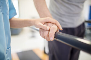 The hand of a young therapist rests on the hand of a senior man as he walks on the parallel board during a physical therapy session.