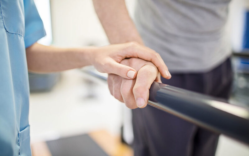 The hand of a young therapist rests on the hand of a senior man as he walks on the parallel board during a physical therapy session.