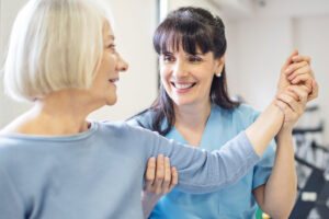 A senior woman with her left arm raised shoulder-height to her side with the help of a younger smiling therapist who has her right hand placed gently on the woman's triceps and her left hand on the woman's wrist.
