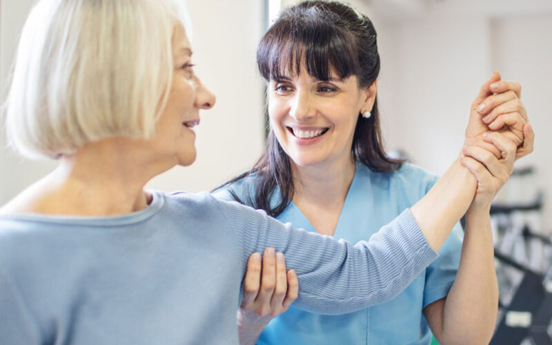 A senior woman with her left arm raised shoulder-height to her side with the help of a younger smiling therapist who has her right hand placed gently on the woman's triceps and her left hand on the woman's wrist.