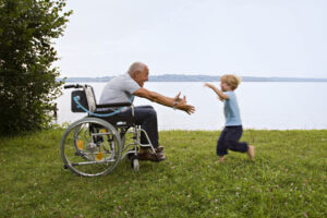 A smiling senior man sitting in a wheelchair with his arms outstretched in front of him to greet a young boy who is enthusiastically running toward the man. The pair are in a grassy field on the edge of a calm lake on a slightly hazy day.