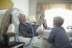 A senior man wearing a respiratory ventilator placed over his nose while lying in a bed in a well-decorated room. A smiling young woman and a younger boy are beside the man, leaning on his bed.